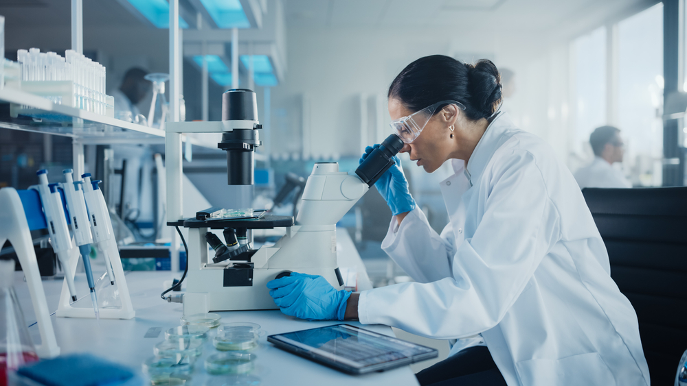 Kvinnelig forsker i et laboratorium, som lener seg over og ser inn i et mikroskop. 
