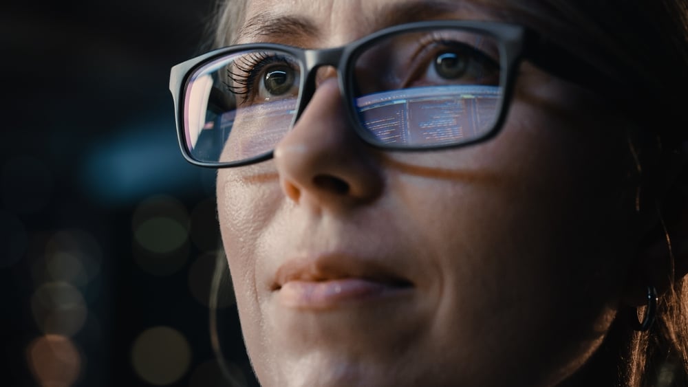 Et nærbilde av en kvinne som skeptisk ser på dataskjermen, hvor skjermen hennes reflekteres gjennom hennes briller..